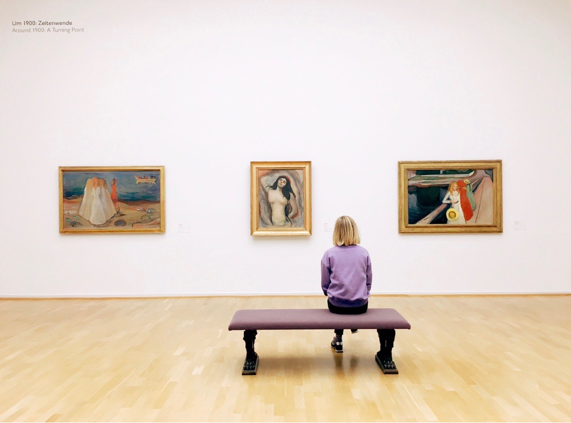 A woman is sitting in an art gallery and looking at three artworks hanging on the wall