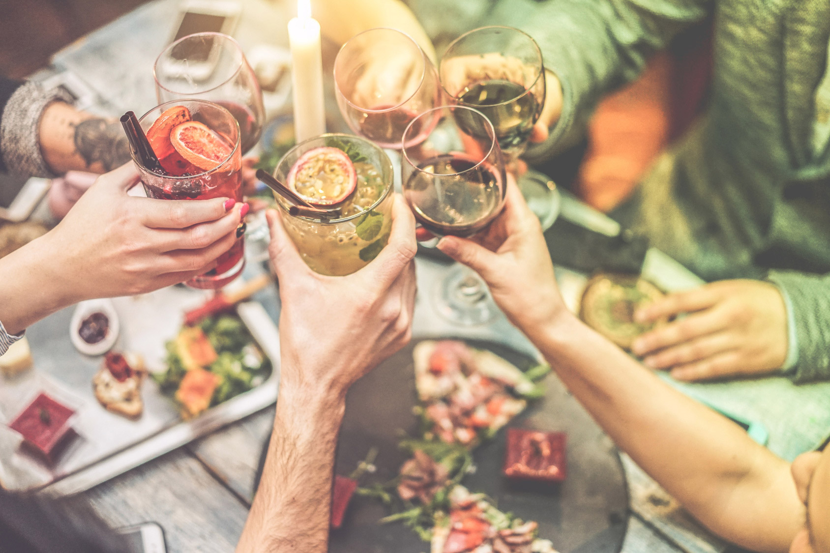 Six people are cheering together with cocktails and wines over a table with food