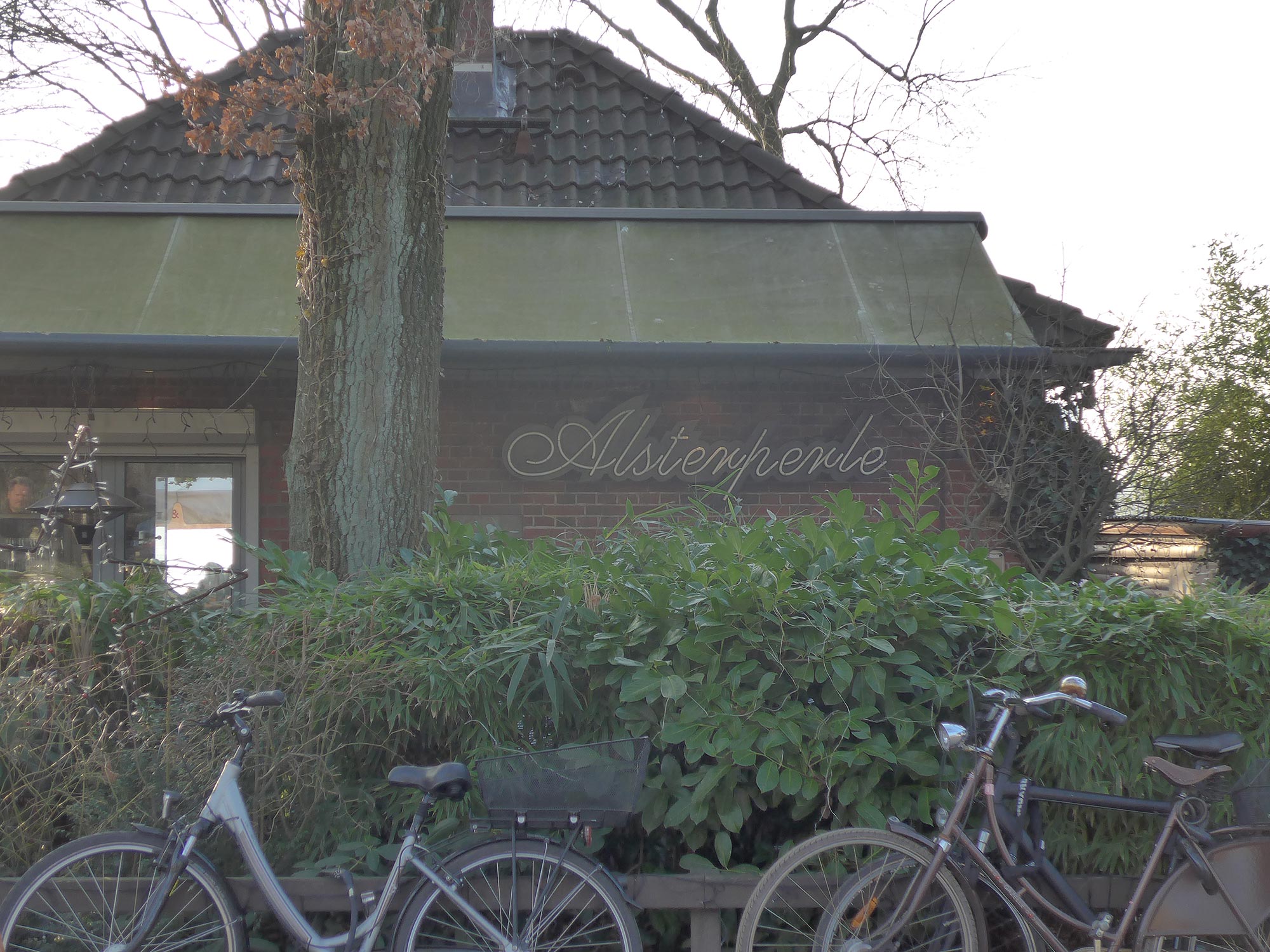 Zwei Fahrräder lehnen an einem Gebüsch vor einem kleinen Haus mit der Aufschrift Alsterperle