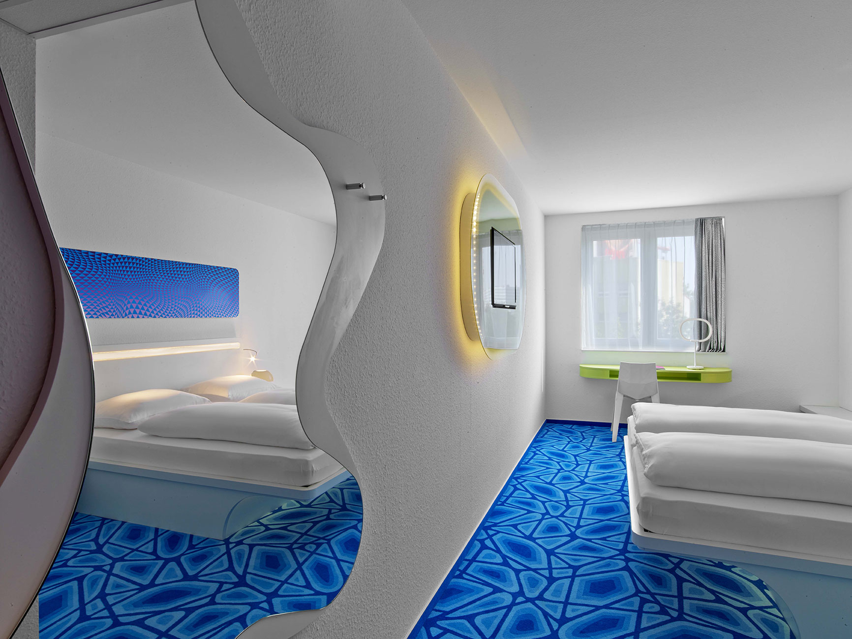Bright hotel room with a blue floor, white double bed and a built-in TV
