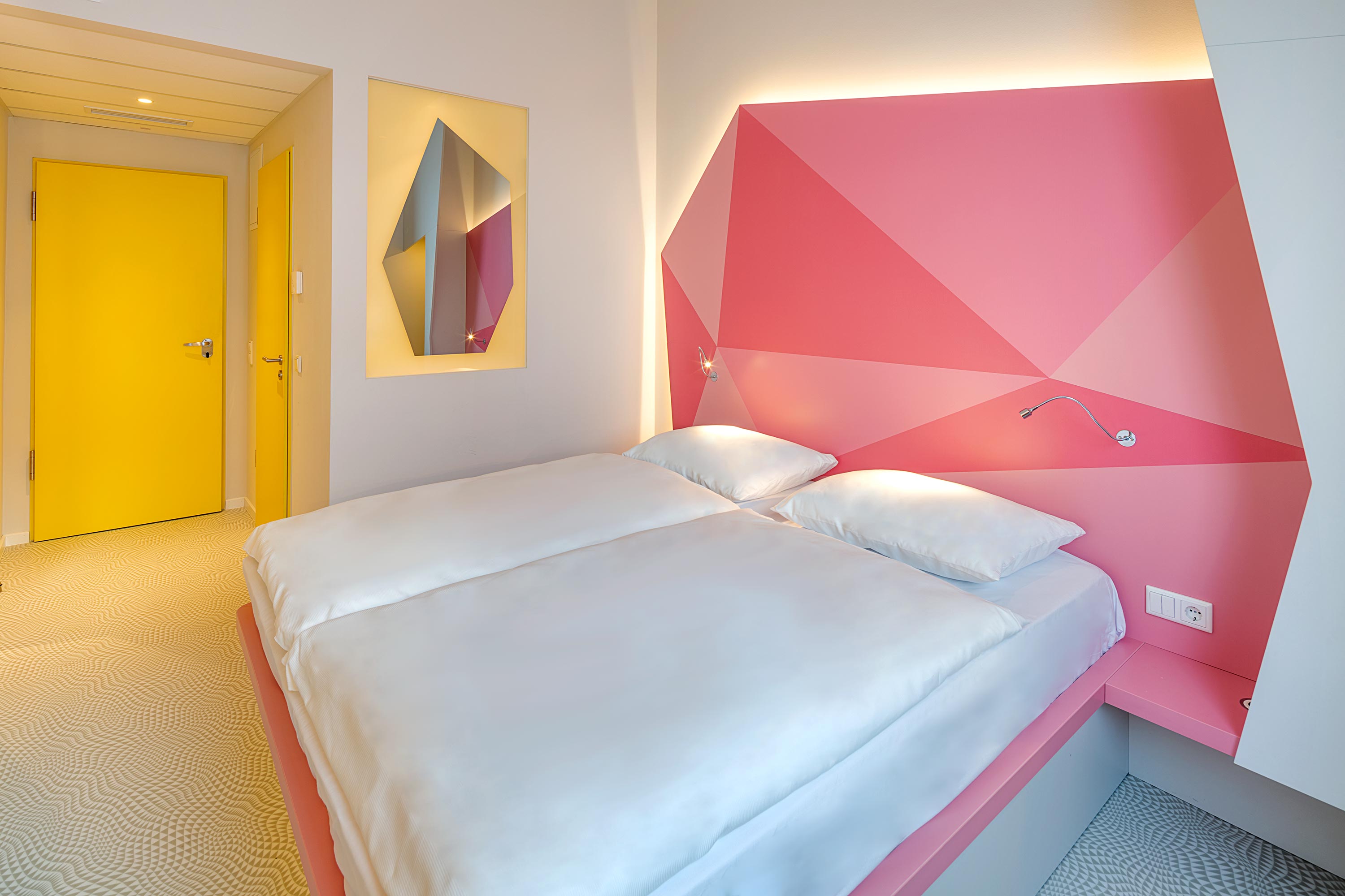 Ein Doppelbett vor einer rosa beleuchteten Wand und einer gelben Türe im Hintergrund