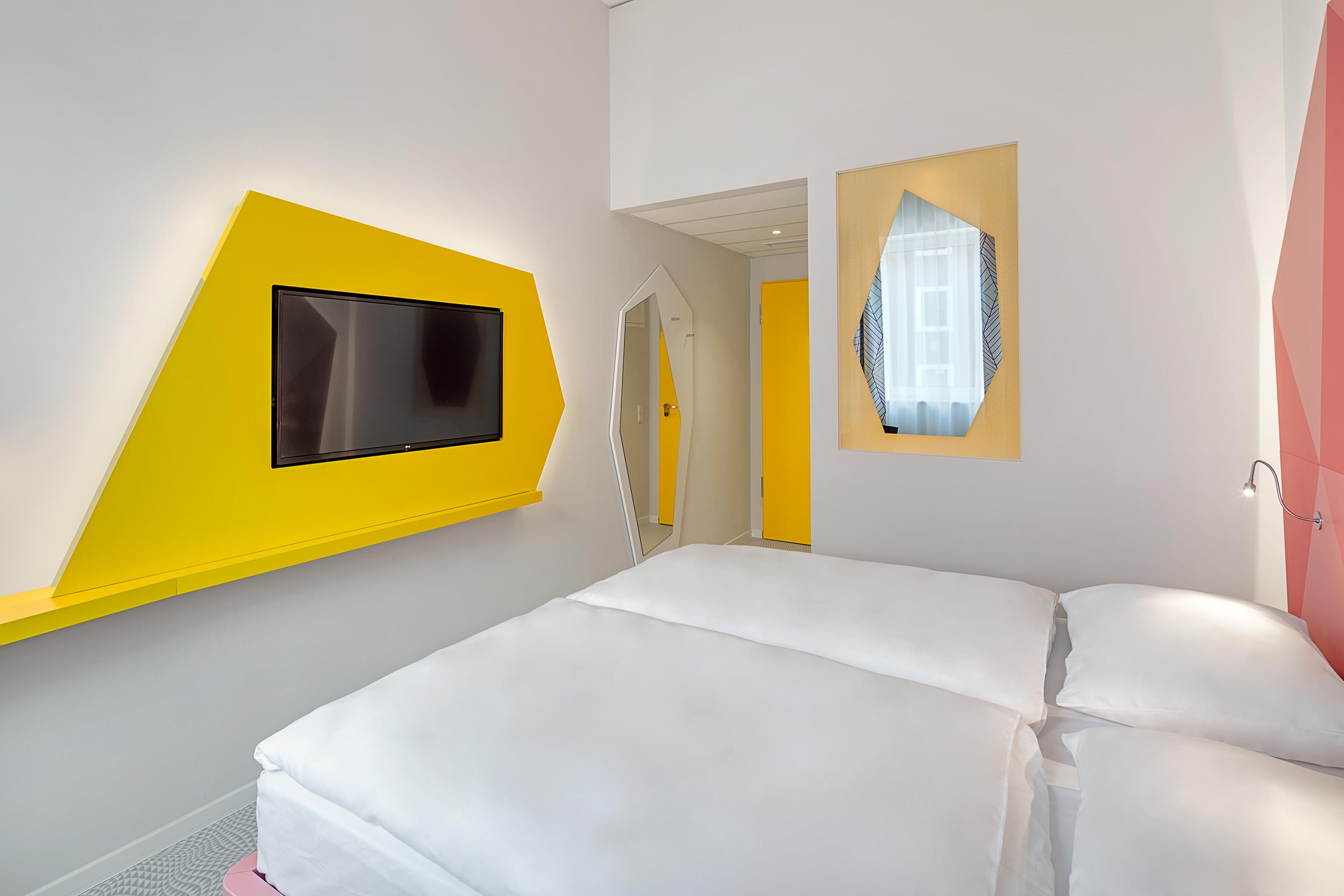 Ein frisch bezogenes Bett vor einem Flachbildfernseher, der an einer gelben Wand hängt